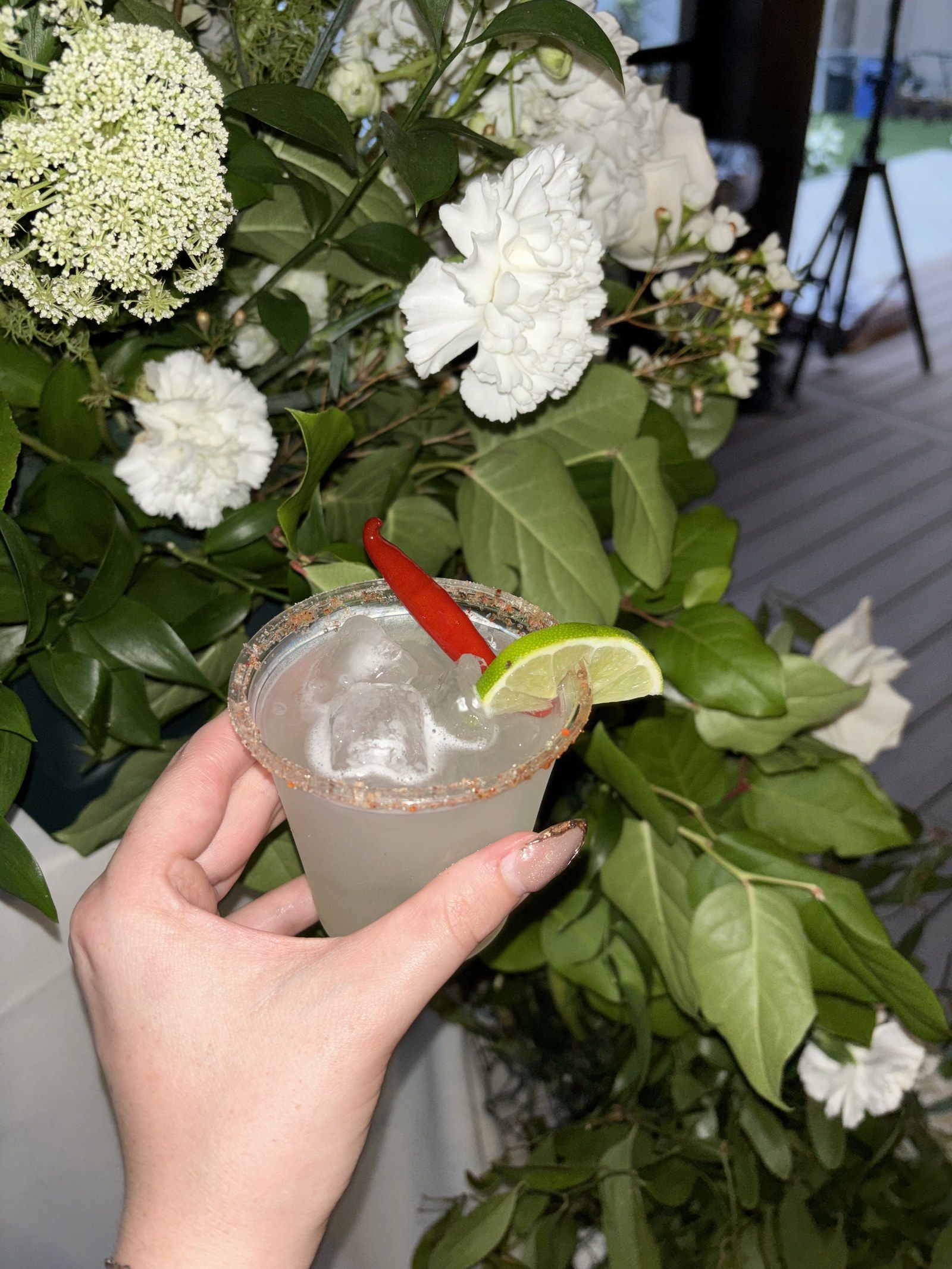 A pale margarita with a chili-salt rim, a red chili and a lime wedge, held up in front of white flowers