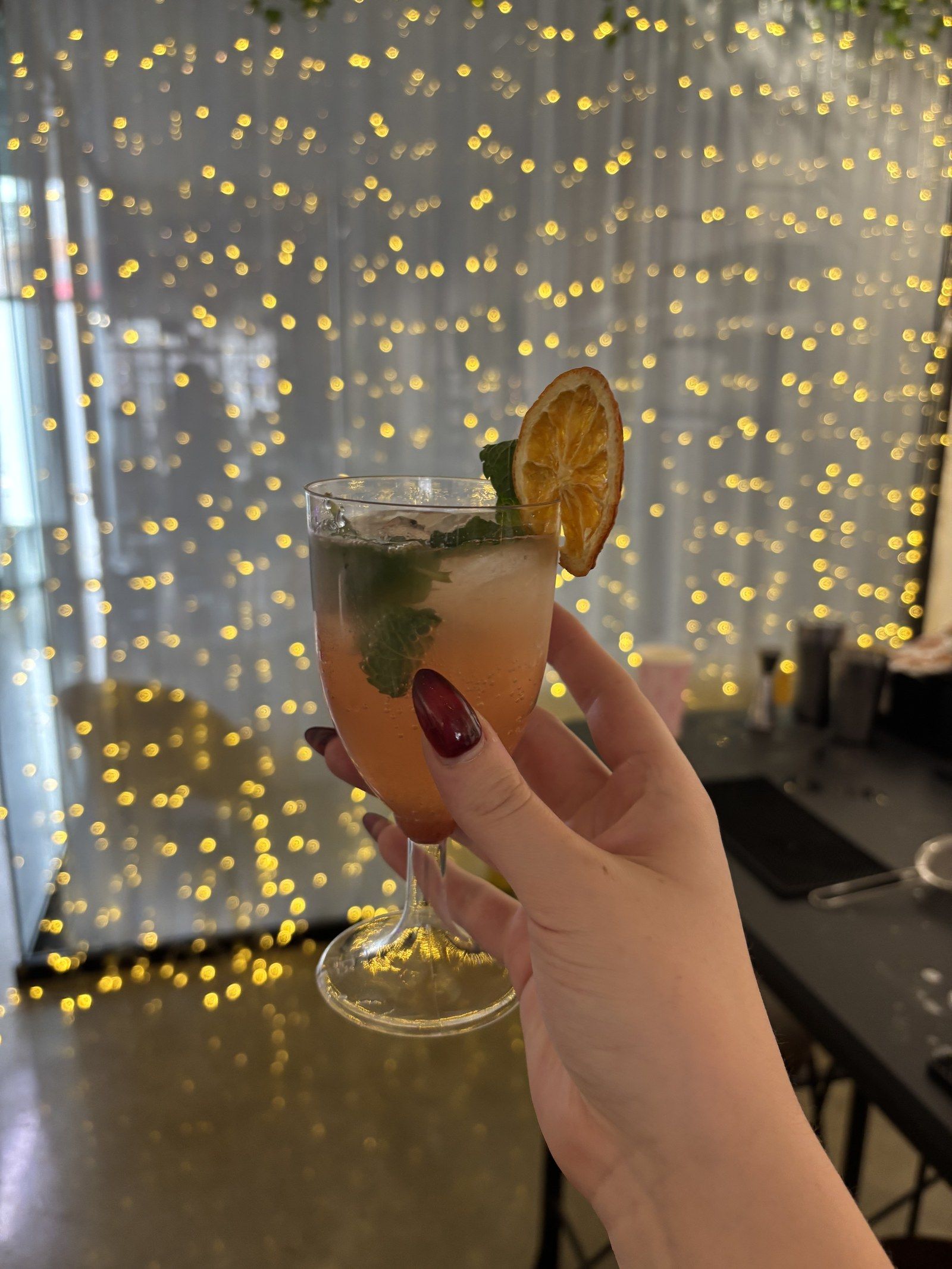 An orange drink with mint and a dried orange wheel, held up in front of a wall of fairy lights