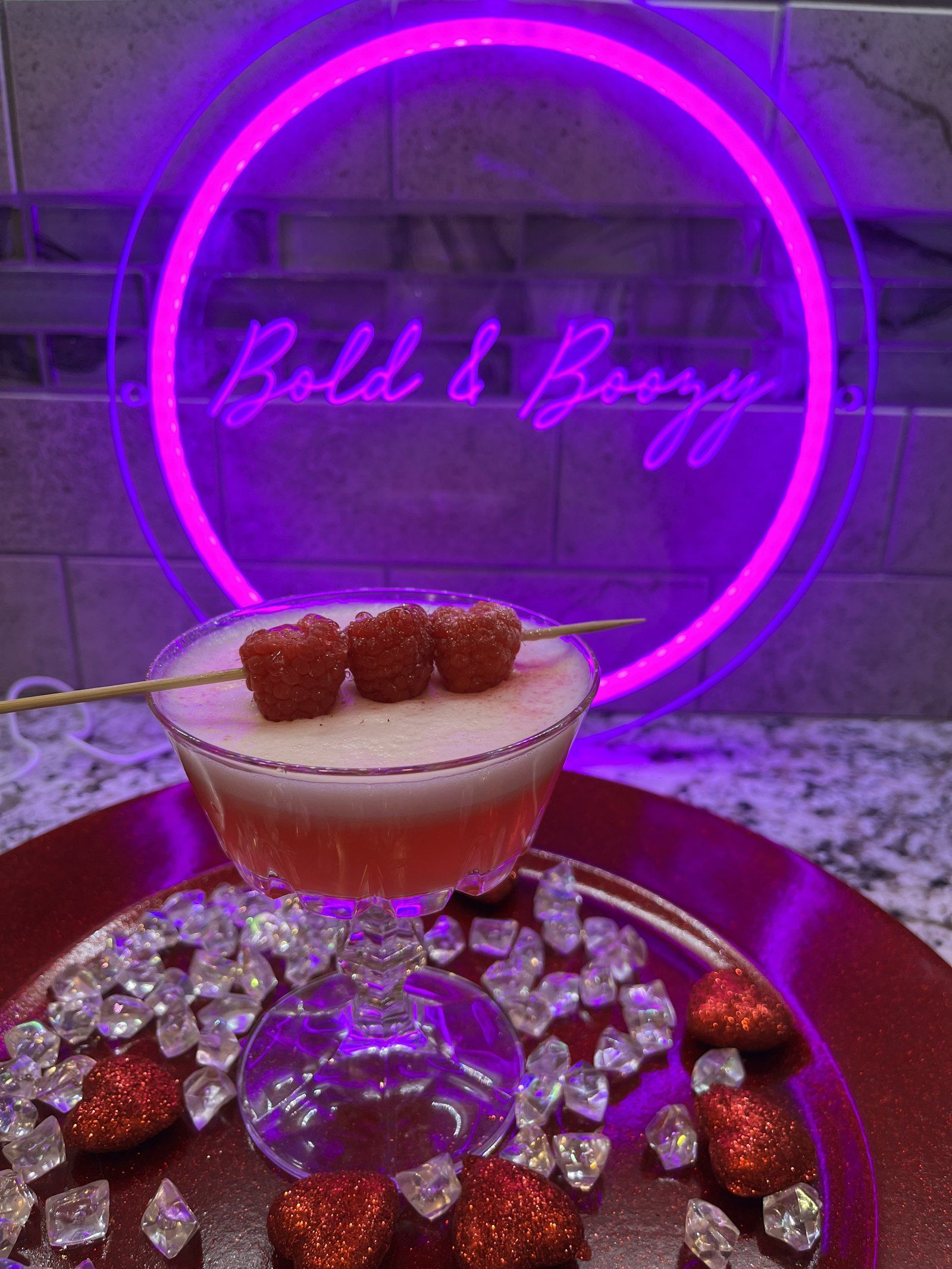 A pink cocktail in a coupe with a skewer of raspberries, on a tray of crystals in front of the glowing Bold & Boozy neon sign