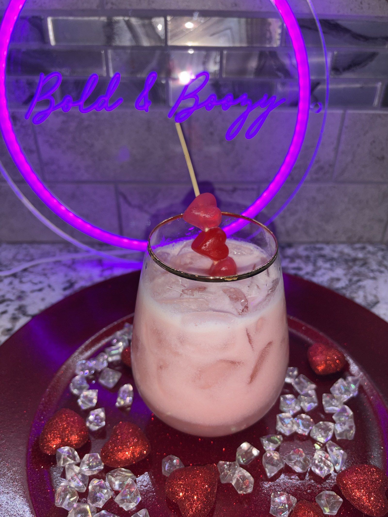A creamy pink drink with a skewer of gummy hearts in front of the Bold & Boozy neon sign